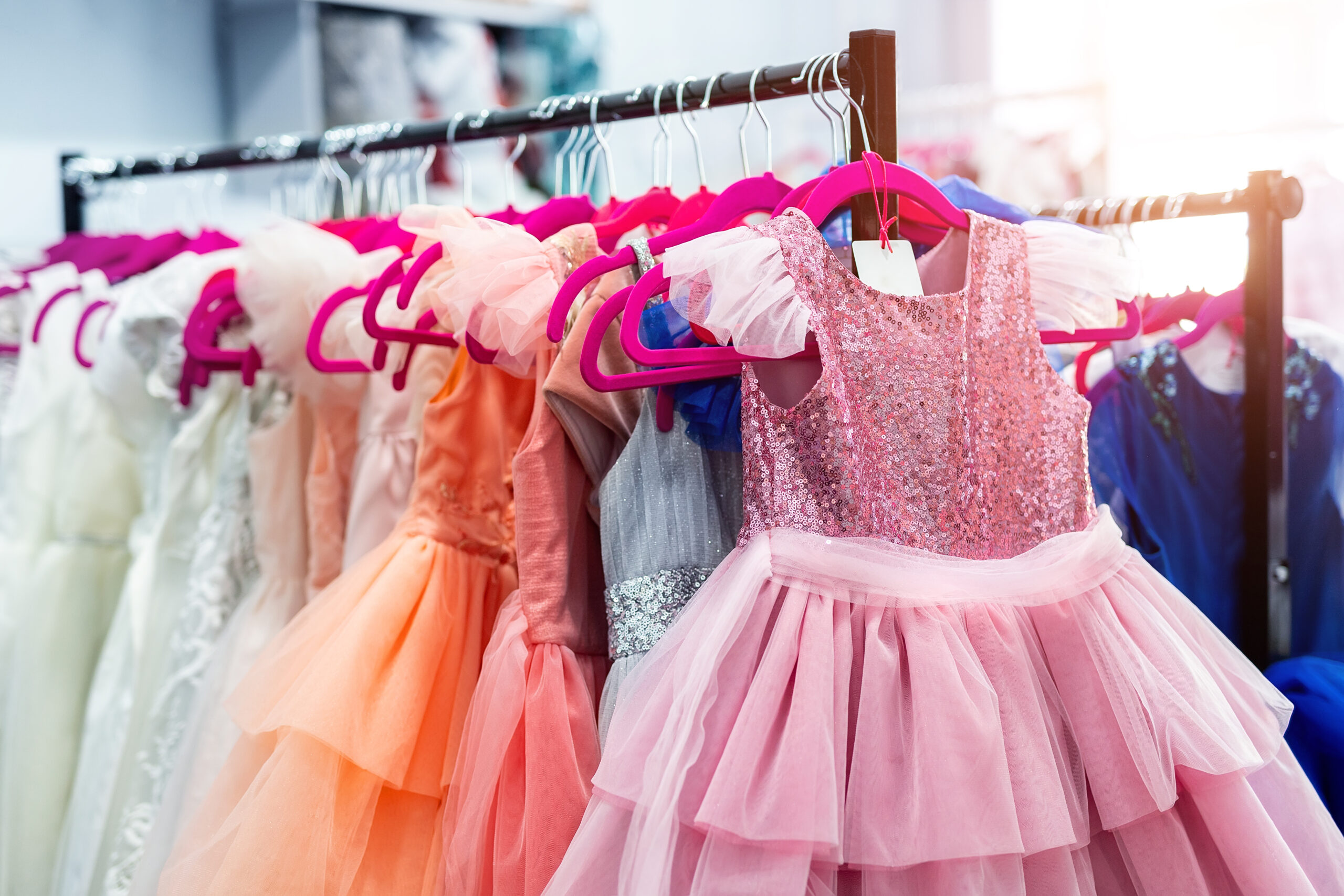Rack with many beautiful holiday dresses apparel on hangers at female fashion showroom indoor. Kid girl dress hire studio for celebration birthday party or photography session event. Bright sun day.